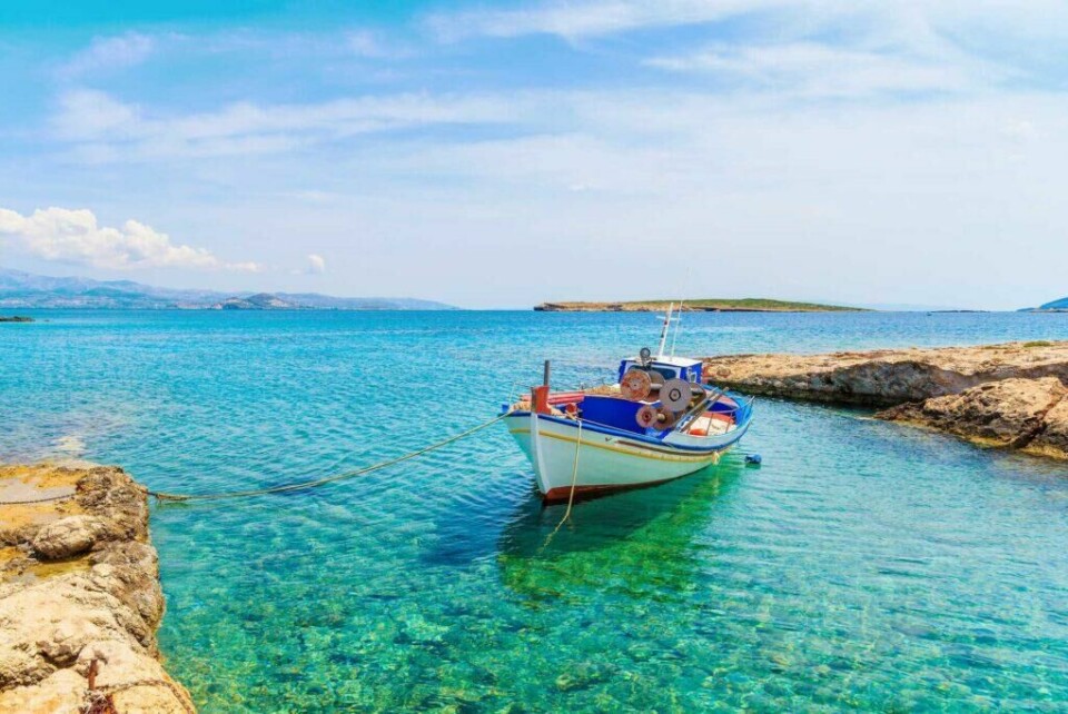 Opplev flere kulturer med øyhopping i Hellas. (Foto: Apollo) Bildet viser en liten tradisjonell fiskebåt som ligger fortøyd i krystallklart turkist vann. Båten er malt i hvitt, blått og rødt, og den ligger i en rolig bukt omgitt av lave klipper. I bakgrunnen kan man se en øy og en blå himmel med lette skyer.