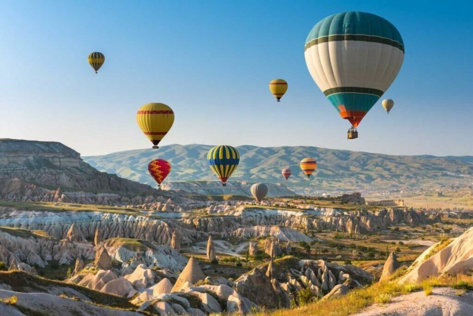 Et fenomen som er på manges liste å oppleve er luftbalingene over Tyrkia. (Foto: Apollo) Bildet viser flere fargerike varmluftsballonger som svever over et unikt landskap med steinformasjoner og daler i Cappadocia, Tyrkia. Landskapet består av myke åser, dramatiske klippeformasjoner og grønne sletter, mens himmelen er klar og blå, og ballongene skaper en magisk stemning.
