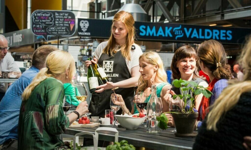 Mathallen Oslo ligger på Vulkan-området – midt mellom øst og vest i Oslo Sentrum – og er et mat-paradis med mer enn 30 spesialbutikker, kafeer og spisesteder. (Foto: CH / VisitNorway) Bilde av servitør og kvinner ved bord med mat på Mathallen på Vulkan-området i Oslo.