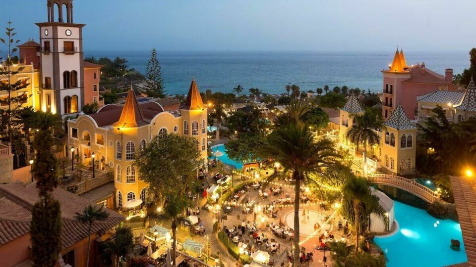 Gran Hotel Bahia del Duque byr på et idyllisk bassengområde omgitt av palmer, grønt og luksuriøse fasiliteter – et av de topp 5 hotellene på Tenerife. Bildet viser en del av det luksuriøse Gran Hotel Bahia del Duque, et av Tenerifes topp 5 hoteller. I forgrunnen er et stort, glitrende basseng med krystallklart vann som reflekterer den blå himmelen. Rundt bassenget står hvite solsenger med tilhørende parasoller, perfekt for avslapning. Høye, slanke palmer strekker seg oppover og gir en tropisk følelse. I bakgrunnen ser man en bygning inspirert av kanarisk arkitektur med små detaljer som gir et snev av eleganse. Atmosfæren er eksklusiv og rolig, som en oase for luksusreisende på Tenerife.
