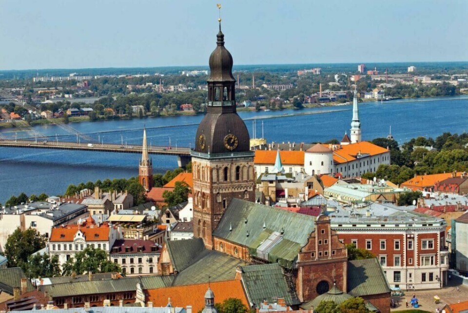 Ta turen til Ringa i 2025! (Foto: Apollo) Bildet viser gamlebyen i Riga, Latvia, med den ikoniske Riga-domkirken i sentrum. Den store, mørke klokkespiren står ut blant bygningene med røde tak. I bakgrunnen renner en bred elv med en moderne bro som forbinder byens to sider. Området rundt er grønt og flatt, med enkelte bygninger i horisonten under en klar, blå himmel.