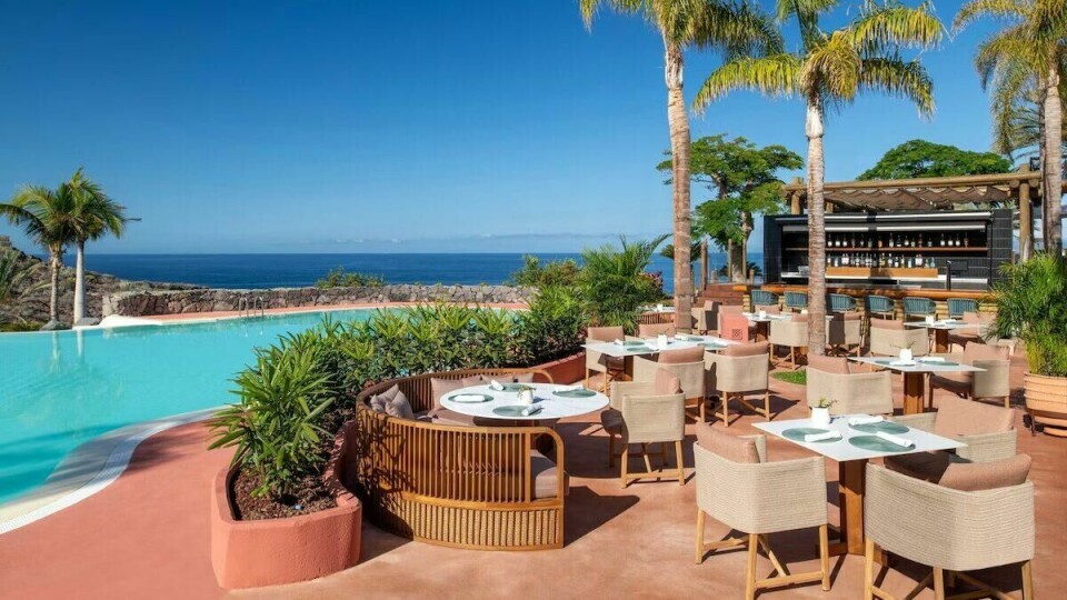The Ritz-Carlton Tenerife, Abama imponerer med sitt vakre bassengområde, frodige hager og eksotisk arkitektur – en ekte perle blant de topp 5 hotellene på Tenerife. Bildet viser det luksuriøse The Ritz-Carlton Tenerife, Abama, et av de topp 5 hotellene på Tenerife. Hotellet er omgitt av vakker natur, med frodige grønne hager, palmetrær og en spektakulær utsikt over havet. I forgrunnen er det et stort, innbydende bassengområde med krystallklart vann, solsenger og parasoller. Hotellets terrakottafargede bygninger har en unik maurisk-inspirert arkitektur som gir en følelse av eksotisk eleganse. Atmosfæren er avslappende og luksuriøs, perfekt for en ferie i rolige og sofistikerte omgivelser.