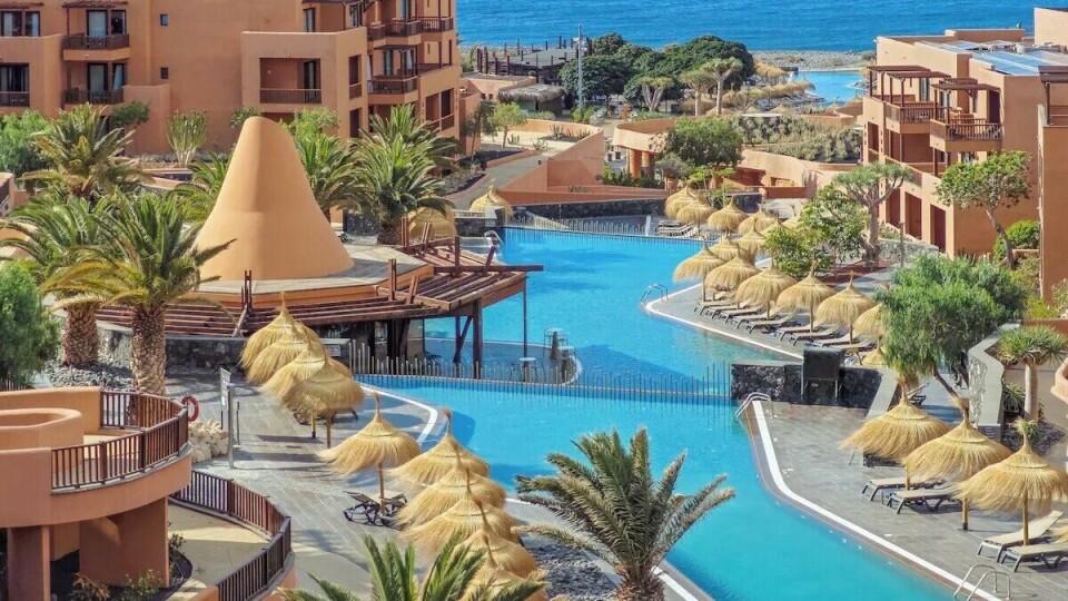 : Sandos San Blas Nature Resort & Golf med basseng, stråparasoller og utsikt mot Atlanterhavet. Bildet viser Sandos San Blas Nature Resort & Golf på Tenerife, et luksuriøst hotell med en innbydende og moderne arkitektur i jordfarger. I forgrunnen strekker et stort, blått basseng seg gjennom området, omgitt av grønne palmer og elegante parasoller laget av strå. Hotellets bygninger, i varme terrakottatoner, omfavner bassenget og har balkonger som gir utsikt mot både havet og de frodige omgivelsene. I bakgrunnen skimtes det dypblå Atlanterhavet som gir en idyllisk ramme rundt anlegget. Atmosfæren er avslappende og eksotisk – perfekt for en ferie på Tenerife.