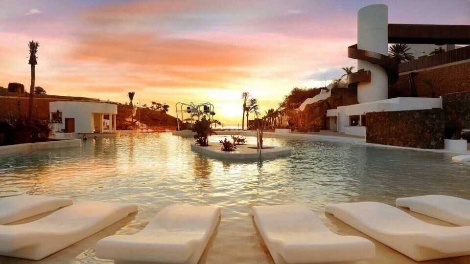 Hard Rock Hotel Tenerife ved solnedgang, med hvite solsenger i vannet og spektakulære oransje og rosa himmeltoner. Et luksuriøst bassengområde omgitt av moderne arkitektur og palmer. Bildet viser et luksuriøst bassengområde ved solnedgang på Hard Rock Hotel Tenerife. Solen går ned i horisonten, og himmelen er fylt med varme oransje, rosa og lilla nyanser som speiler seg i det rolige vannet. I forgrunnen står flere moderne, hvite solsenger delvis nedsenket i vannet, noe som gir en følelse av luksus og avslapning. Midt i bassenget er det små, grønne planter på en øy, og i bakgrunnen ser vi hvite bygninger med moderne arkitektur, palmer og en spiralformet struktur. Hele scenen oser av eleganse og ro.