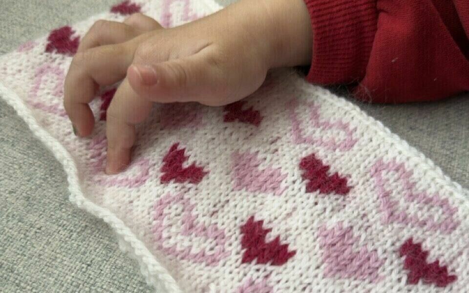 En liten hånd utforsker et strikkemønster med hjerter En barnehånd kledd i en rød genser hviler på et strikket stykke med et mønster av rosa og røde hjerter på hvit bakgrunn. Bakgrunnen består av et lysegrått stoff. Detaljene i strikkearbeidet viser tydelig den myke teksturen i garnet.