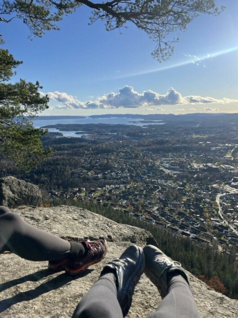 Tar du turen opp til Kolsåstoppene er det stor sjanse for en god tur, nydelig utsikt og en dukkert om du føler for det. To personer sitter på en fjellhylle med bena hvilende ut mot kanten, iført treningstøy og tursko. Under dem brer det seg ut en imponerende panoramautsikt over et tett bebygd område omgitt av skog, med Oslofjorden og flere øyer i det fjerne. Solen skinner fra en blå himmel med noen dramatiske, hvite skyer. Furugrener rammer inn bildet fra toppen, og skaper en naturnær ramme rundt det storslåtte landskapet. Bildet formidler ro, frihet og mestring etter en topptur. Skal du på fjelltur i ferien? Fra Galdhøpiggen til Lofoten; Her har du 10 tips til skjulte turperler hvor du slipper å gå i kø.