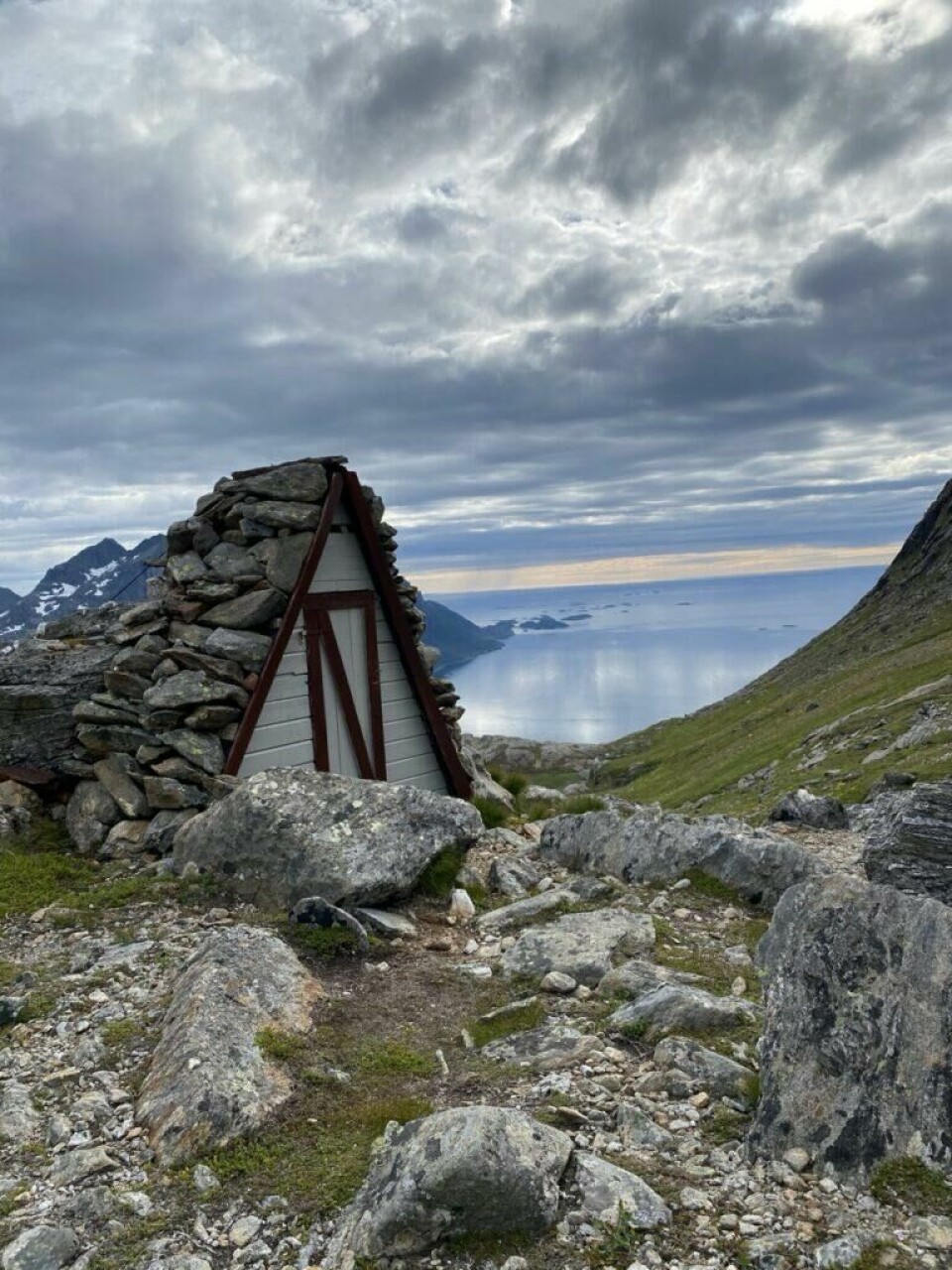 Bilde av toalettet til Skarbua i Skarbotn med utsikt til Brennviksanden i bakgrunnen. En fantastisk flott tur i Steigen i Norland. (Foto: Monika Myhre / ABC Startsiden) Et lite, trekantet bygg med steinvegger og rødmalt trekantdør står midt i et steinete høyfjellslandskap med spektakulær utsikt. I bakgrunnen skimtes havet og øyer langt der nede, badet i et mykt, blått lys under en dramatisk og delvis skydekt himmel. Bratte fjellsider, snøkledde topper og grønne fjellskrenter omkranser hytta, og skaper en følelse av både isolasjon og ro midt i naturens storhet. Bildet formidler ekte villmark og det nordnorske kystlandskapets rå skjønnhet – fra Steigen.Bilde av toalettet til Skarbua i Skarbotn med utsikt til Brennviksanden i bakgrunnen. En fantastisk flott tur i Steigen i Norland.