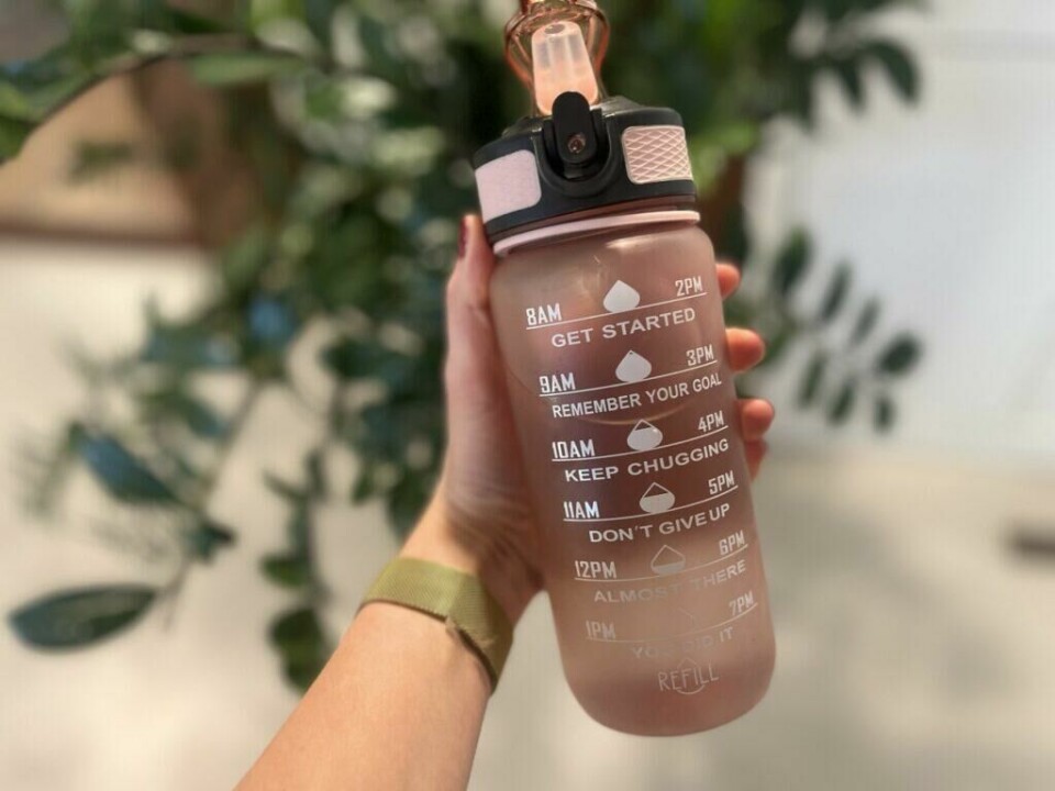 – Drikk mer vann - og gå ned i vekt, skriver ekspertene og forklarer hvordan det gjøres. Vi forteller! (Foto: Cecilie Aasland / ABC Startsiden) Bilde av en hand som holder en rosa motivasjonsflaske foran en grønn plante. Hun har på seg en svart genser og en apple watch i gull, og står foran et vindu. Vi forteller om «vanntabben» som kan send deg opp i vekt! Og forteller hvordan det å drikke vann riktig eller feil kan ha innvirkning på forbrenningen din.