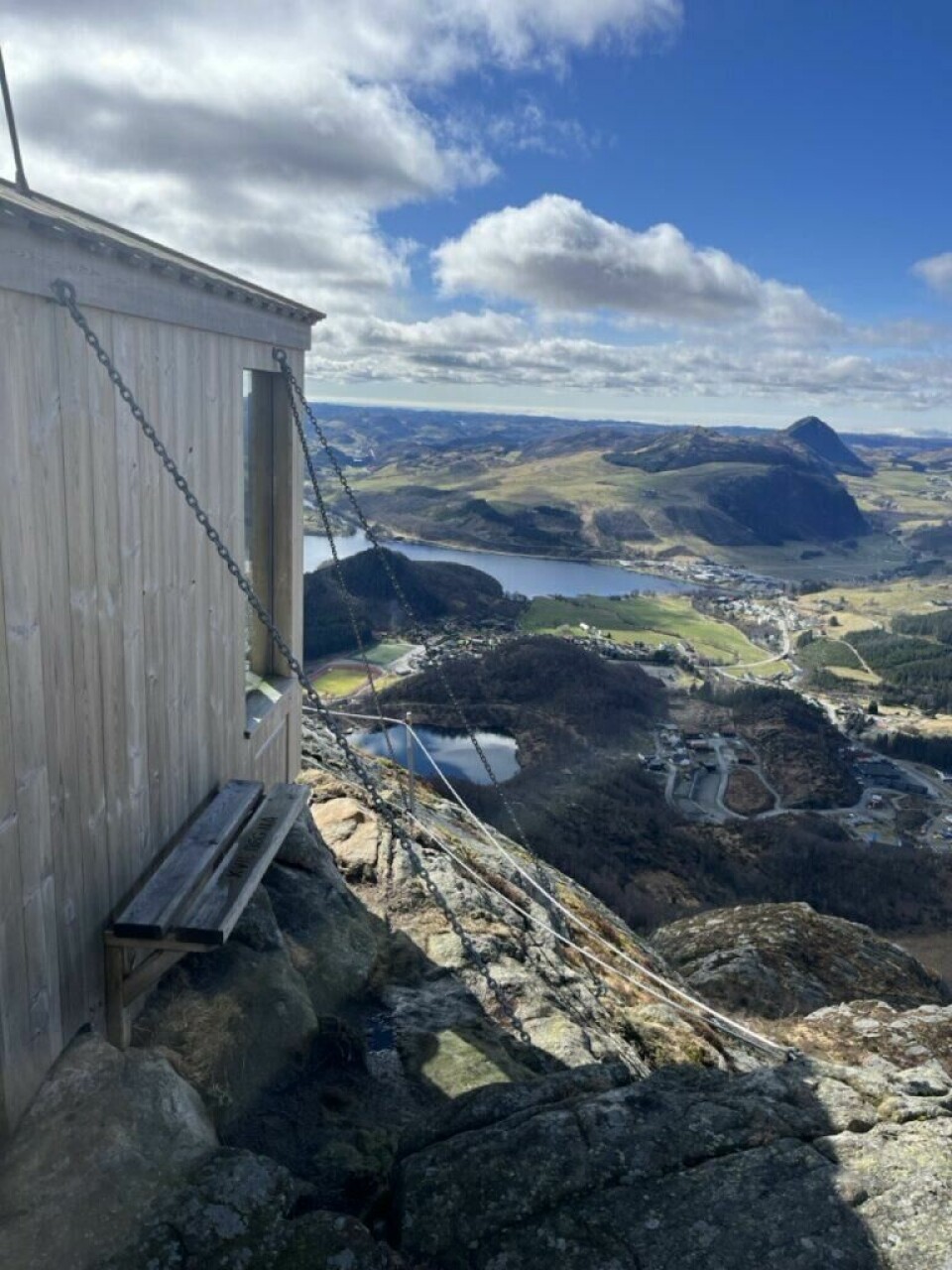 Fra toppen av Storafjellet har du en imponerende utsikt og du ser store deler av Dalane, til havet på Jæren og til tettstedet Ålgård. (Foto: Cecilie Aasland / ABC Startsiden) Et fjellandskap åpner seg i dramatisk utsikt fra en gapahuk som er plassert helt på kanten av et stup. Gapahuken er solid festet med kraftige kjettinger og har en enkel benk foran seg med teksten 'TINDEGRISAN' inngravert. Utsikten strekker seg utover Vikeså i Bjerkreim, med åser, vann og landbruksområder badet i sollys under en blå himmel med spredte skyer. Et markant, spisst fjell sees i bakgrunnen, og nedenfor fjellsiden ligger bebyggelse og veier godt synlig. Bildet formidler følelsen av frihet, mestring og storslått norsk natur.Fra toppen av Storafjellet har du en imponerende utsikt og du ser store deler av Dalane, til havet på Jæren og til tettstedet Ålgård.