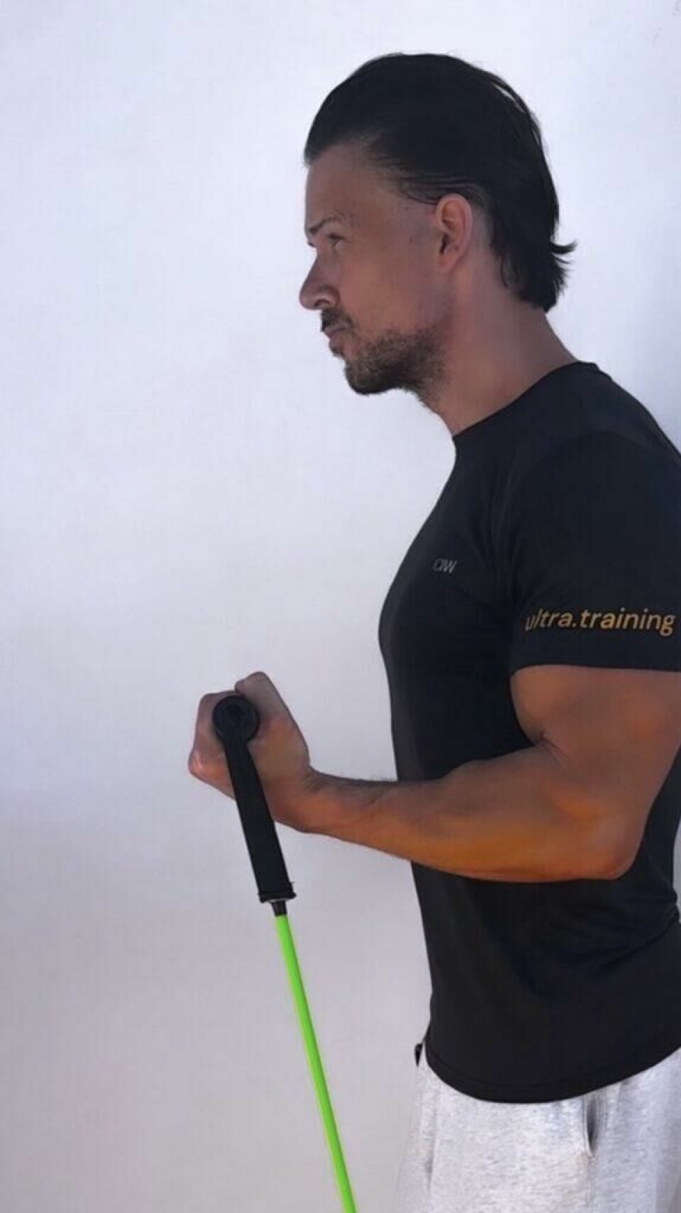 Bicep curl: Hold enden på en treningsstrikk i hver hand. Bøy så albuen og løft manualen opp mot skulderen til håndflaten din er vendt inn mot skulderen din. Bilde av personlig trener Cornelis Elander som trener bicep curl stående og med treningsstrikk. Dette er en øvelse som trener biceps og den kan gjennomføres både stående og sittende, og med treningsstrikk og med manualer. Han har på seg en svart t-skjorte. Dette er en styrkeøvelse med fordeler for armer og biceps, og en av styrkeøvelsene i vårt 30-dagers gratis treningsprogram og styrkeprogram.