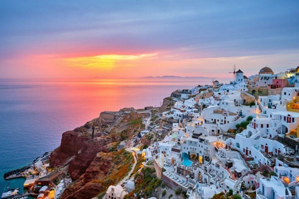 Øyhopping i Hellas: Santorini tilbyr gresk kultur, vakker utsikt og spektakulære solnedganger. Ikke rart byen er så populær blant turister! (Foto: Colourbox) Oversiktsbilde av hvite murhus med blå tak i Santorini, i bakgrunnen ser man en solnedgang som farger himmelen rosa, lilla og orange. Her får du en utfyllende guide på øyhopping i Hellas hvor vi gir deg tre reiseruter, pakkeliste og gode tips til hvordan man gjør øyhopping med barn. Opplev plasser som Santorini, Kreta, Athen, Paros, Naxos, Korfu og Mykonos.