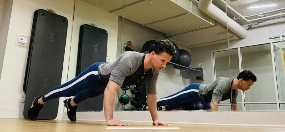 Push-ups med bredt grep: En riktig utført armheving starter med en korrekt «planke», og det skal mer eller mindre gå en rett linje fra topp til tå. (Foto: Cornelis Elander) Bilde av personlig trener Cornelis Elander som trener armhevinger på treningsmatte. Han har på seg en grå t-skjorte, en blå treningsbukse og befinner seg i et treningsrom. Dette er en styrkeøvelse med fordeler for hele kroppen, og er en av styrkeøvelsene i vårt 30-dagers gratis treningsprogram og styrkeprogram.
