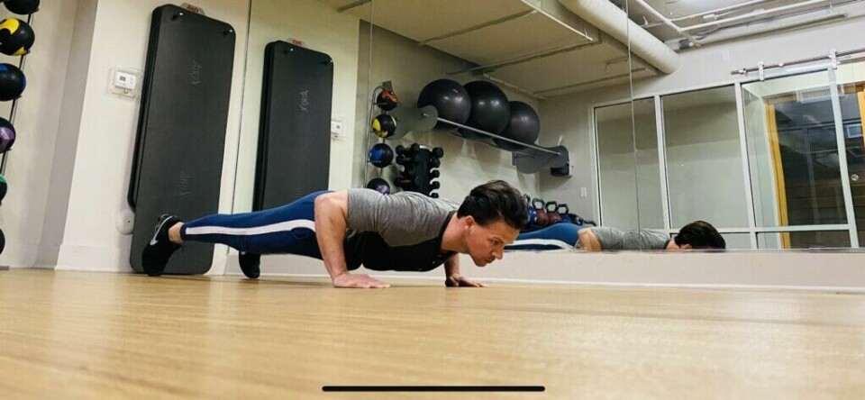 Pushups med bredt grep: Det er kun brystkassen som så vidt kan berøre underlaget, mens hofte, midje, knær, og resten av kroppen IKKE skal kollapse ned mot underlaget eller forandre posisjon underveis. (Foto: Cornelis Elander) Bilde av personlig trener Cornelis Elander som trener armhevinger på treningsmatte. Han har på seg en grå t-skjorte, en blå treningsbukse og befinner seg i et treningsrom. Dette er en styrkeøvelse med fordeler for hele kroppen, og er en av styrkeøvelsene i vårt 30-dagers gratis treningsprogram og styrkeprogram.