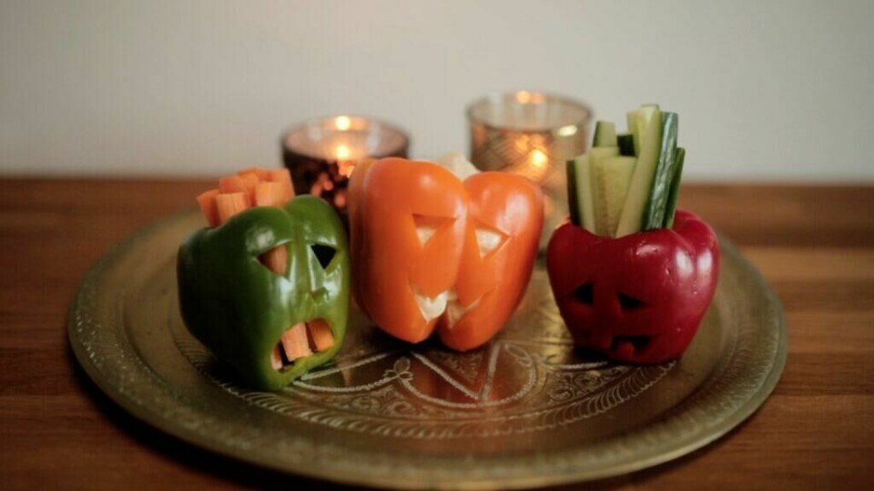 Paprikaer skåret ut som halloween-lykter, fylt med friske grønnsaksstaver og dip. (Foto: Kreative ideer/ Aurora Lange) Bildet viser tre paprikaer i grønt, oransje og rødt, som er skåret ut med ansikter som ligner tradisjonelle halloween-gresskar. Paprikaene er fylt med grønnsaksstaver som gulrøtter og agurker, og den oransje paprikaen ser ut til å inneholde en dip. Paprikaene står på et dekorativt metallbrett, og i bakgrunnen er tre tente telysholdere som gir et varmt lys. Dette er en morsom og kreativ måte å servere snacks på til en halloweenfest.