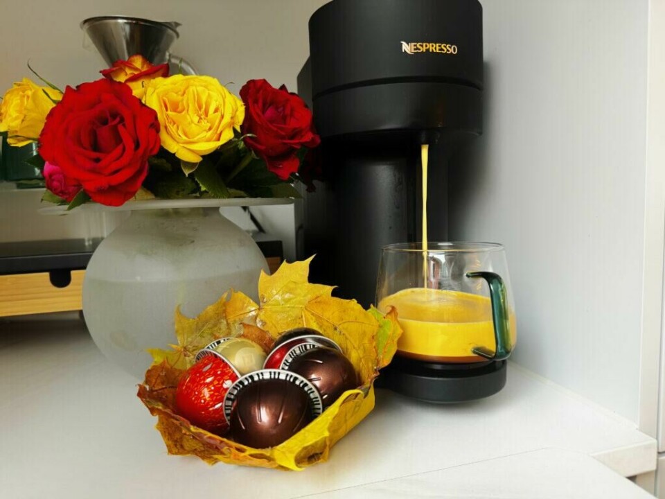 Kaffehjørne med vakre roser og en hjemmelaget kaffekapselskål av blader. Bildet viser en Nespresso kaffemaskin i aksjon, som lager en kopp kaffe som helles i et glasskrus. Ved siden av kaffemaskinen står en vase med friske roser i røde og gule nyanser, noe som gir et livlig og fargerikt preg til scenen. Foran vasen er en liten skål laget av høstblader, som holder flere kaffekapsler i ulike farger. Denne kreative skålen tilfører et naturlig og unikt element til kaffehjørnet, som kombinerer praktisk funksjon med estetisk dekorasjon.