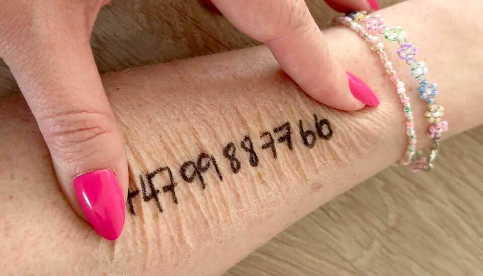 Skriv telefonnummeret tydelig på barnets arm med hudvennlig tusj før du beskytter det med plasterspray. Et nærbilde av en voksen hånd som peker på et telefonnummer skrevet med svart eyeliner på underarmen til et barn. Barnet har på seg perlearmbånd i pastellfarger. Huden er synlig tørr, og nummeret er tydelig skrevet på huden. Bildet viser hvordan man kan skrive kontaktinformasjon direkte på barnets hud før man påfører plasterspray.
