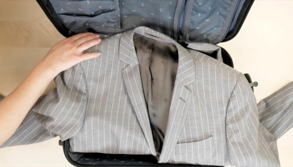 Legg dressjakken flatt i kofferten før du pakker resten. Brett den til slutt over innholdet for å unngå skrukker. Åpen koffert med en grå stripete dressjakke lagt flatt over bunnen. En hånd justerer jakken forsiktig før resten av bagasjen pakkes oppå. Dette er en metode for å unngå skrukker i dressjakken under reise.