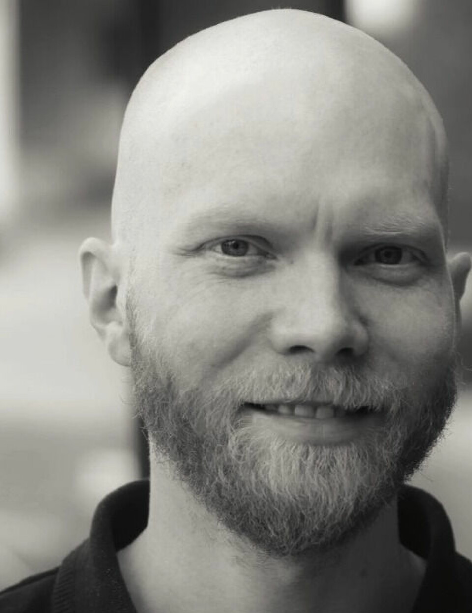 Ole Jenssen er daglig leder for Oxylab, og har testet Ole Aaknes sin VO2-maks. Portrett av en mann med glattbarbert hode og lys skjeggvekst. Han ser rett mot kameraet og smiler svakt. Bildet er i gråtoner, og bakgrunnen er uskarp, noe som gir motivet god dybde og fokus.