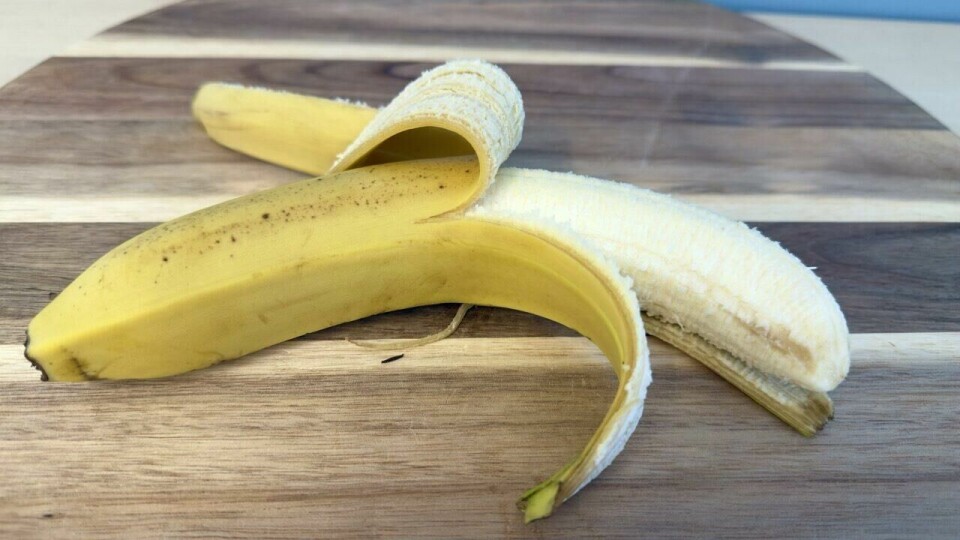 En moden banan, delvis skrelt, plassert på et stilrent skjærebrett – perfekt for et sunt mellommåltid. En delvis skrelt banan ligger på et skjærebrett av tre med mørke og lyse striper. Den gule skallet er bøyd tilbake, og den modne, lyse frukten er tydelig synlig. Bakgrunnen har en lys blå fargetone, som gir en frisk og innbydende kontrast.