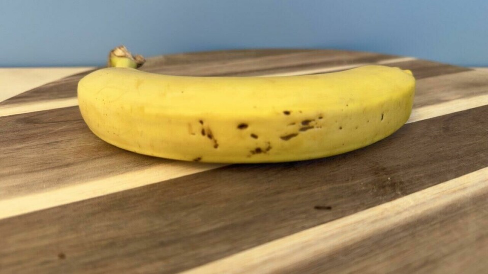 En fersk banan med et naturlig modnet skall, klar til å nytes som en sunn snack. En moden gul banan med små brune flekker ligger på et stilrent skjærebrett av tre med lyse og mørke striper. Bakgrunnen har en lys blå fargetone, som skaper en behagelig kontrast til frukten.