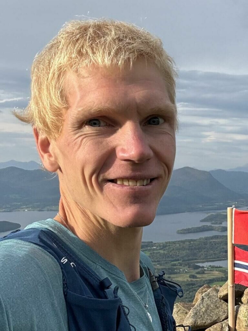 Torbjørn Høstmark Borge står bak nettsiden Pulssonen.no. En smilende mann, Torbjørn Høstmark Borge fra pulssonen om treningsklokker, tar en selfie på toppen av et fjell med vidstrakt utsikt over fjorder, øyer og fjell i bakgrunnen. Han har lyst, kort hår og er iført en blå teknisk t-skjorte med en blå løpevest (med påskriften '12 ADV SKIN'). Bak ham ser vi en varde bygget av steiner med en norsk flaggstang stikkende opp, hvor et slitt norsk flagg og et rødt flagg med et ukjent emblem vaier i vinden. Været er overskyet, men med noen solgløtt som gir god sikt over landskapet. Langt der nede sees tettbebyggelse, grønne jorder og vannveier som slynger seg mellom øyene. Den dramatiske, men fredelige naturen antyder at dette er et populært turmål i Norge.