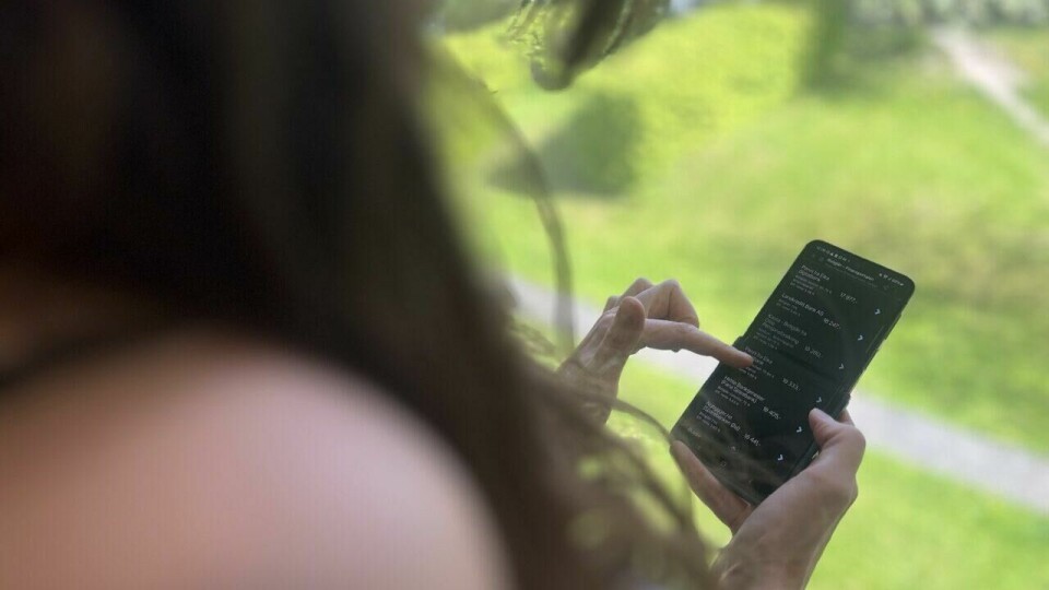 Blakk med god lønn? Her er strategien som virker – steg for steg En person står ved et vindu med utsikt over en grønn park eller hage. Personen holder en smarttelefon i hånden og bruker pekefingeren til å navigere i en app med mørk bakgrunn og lys tekst. Skjermen viser en liste over transaksjoner eller kontoinformasjon. Fokus er på telefonens skjerm og personens hånd, mens bakgrunnen og personen selv er delvis ute av fokus. Lyset fra utsiden gjør at bildet får et lyst og sommerlig preg.