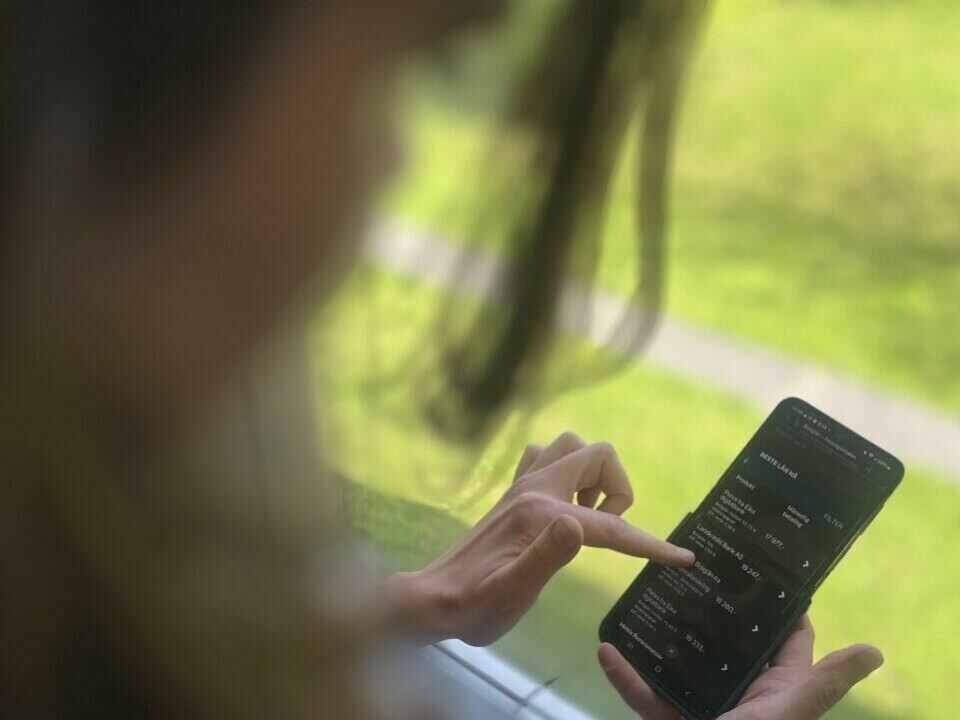 Olaussen anbefaler å ta en ting av gangen og å ikke gape over for mye på én gang. – Jeg prøvde selv veldig mye samtidig, og ville både betale ned gjeld, spare til ferie, buffer – og da gikk jeg på en smell igjen. En person står ved et vindu med utsikt over en grønn park eller hage. Personen holder en smarttelefon i hånden og bruker pekefingeren til å navigere i en app med mørk bakgrunn og lys tekst. Skjermen viser en liste over transaksjoner eller kontoinformasjon. Fokus er på telefonens skjerm og personens hånd, mens bakgrunnen og personen selv er delvis ute av fokus. Lyset fra utsiden gjør at bildet får et lyst og sommerlig preg.