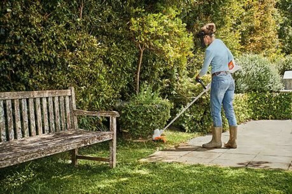 En batteridrevet kantklipper er gjerne stillegående og effektiv, så er lydnivå avgjørende for deg kan dette være riktig valg. (Foto: Garden Store) Kvinne som står med kantklipper/ gresstrimmer i dongeribukse i hage og klipper kant rundt busk.