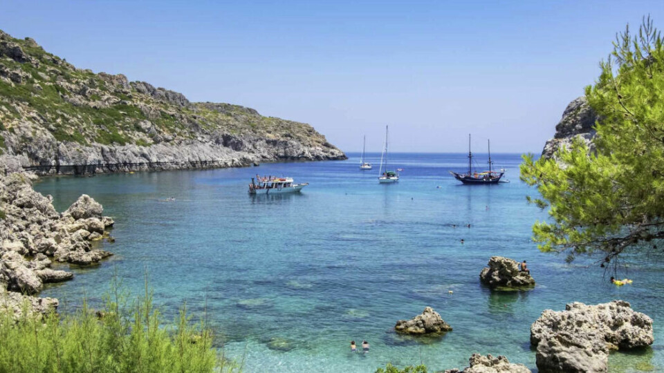 Krystallklart badevann og sjarmerende båtbukt på Rhodos – en av sommerens mest solsikre restplass-destinasjoner i juni. Bildet viser en vakker, beskyttet bukt omgitt av klipper og grønn vegetasjon på Rhodos. Det klare, turkise vannet er stille og innbydende, og flere små båter og seilskuter ligger for anker ute i bukten. Nær land svømmer folk i det grunne vannet, mens noen sitter på steiner som stikker opp fra sjøen. I forgrunnen ses busker og furutrær som rammer inn utsikten mot det glitrende havet. Alt oser av avslappet ferieidyll i en rolig, naturlig setting.