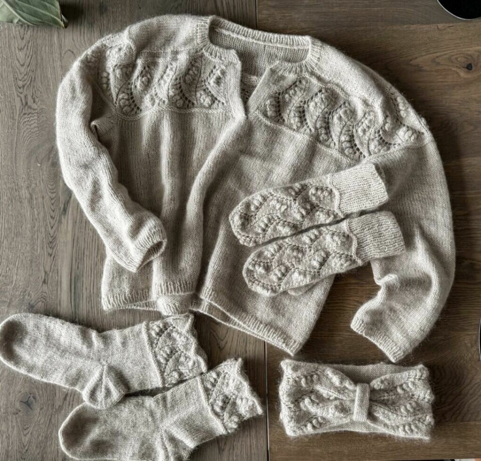 Hele Bluebell-serien: genser, votter, sokker og pannebånd – alt strikket med samme elegante mønster. Flatlay av Bluebell-serien fra Mine Gleder, som inkluderer Bluebell Sweater, votter, sokker og pannebånd. Alle delene er strikket i en lys ullblanding og pyntet med det karakteristiske popcorn- og bladmønsteret. Plaggene er plassert på et bord og viser helheten i designet.