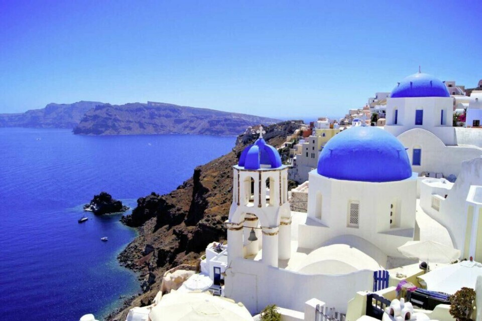 Santorini, med sine ikoniske hvite hus med blå tak, er en av våre anbefalte reisemål hvor du kan sikre deg billige reiser og ferier med restplass. (Foto: Apollo) Bildet viser den ikoniske utsikten fra Santorini, Hellas, med de karakteristiske hvite bygningene og blå kuplene i forgrunnen. I bakgrunnen strekker det dype blå Egeerhavet seg mot horisonten, med en fjellrik øyformasjon i det fjerne. De hvitkalkede bygningene klamrer seg til klippene og skaper en spektakulær kontrast til det asurblå havet og himmelen. Bildet fanger den typiske arkitekturen og den idylliske atmosfæren som gjør Santorini til en av de mest ettertraktede reisemålene i verden. Dette er et perfekt postkortmotiv for reiselystne, med sin kombinasjon av vakker natur, unike bygninger og en følelse av ro og eksklusivitet. Drømmer du om Rhodos og Santorini? Nå kan du oppleve den greske drømmen til en billig penge. Her finner du restplasser til Hellas!