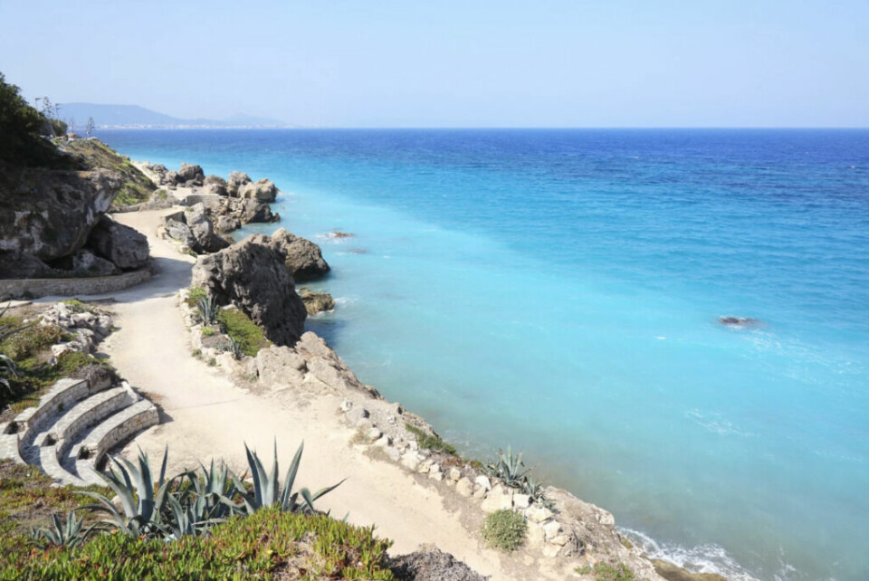 Rhodos er den varmeste øya i Hellas med en gjennomsnittstemperatur på 24 grader, her har du svar på 10 ofte stilte spørsmål om Hellas. (Foto: Canva) Bildet viser en vakker kystlinje med turkist, krystallklart vann som skifter fra lys blå nær stranden til en dypere blåfarge lenger ut. Langs kysten går en svingete sti omgitt av klipper og frodig, grønn vegetasjon, inkludert sukkulenter og agaveplanter. Stien ser ut til å være en populær turvei med en liten steinbenk eller utkikkspunkt på venstre side. I bakgrunnen kan man se en fjern kyststripe med hvite bygninger, som antyder at bildet er tatt fra en av Hellas’ øyer, sannsynligvis Rhodos, kjent for sitt varme klima og idylliske strender. Dette bildet formidler en følelse av ro, naturskjønnhet og eventyr – perfekt for reisende som ønsker å oppleve Hellas’ kystlandskap, enten til fots eller for en avslappende ferie ved havet. Drømmer du om Rhodos, Samos og Santorini? Nå kan du oppleve den greske drømmen til en billig penge. Her finner du restplasser til Hellas!